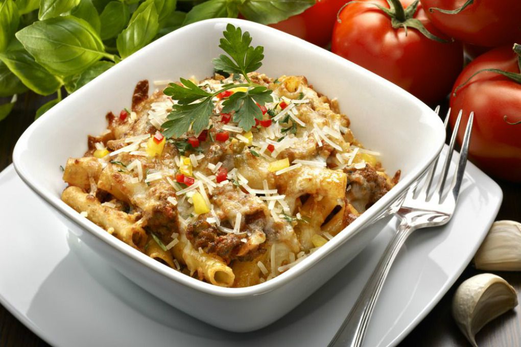 A bowl of beefy macaroni and cheese with whole tomatoes and basil in the background