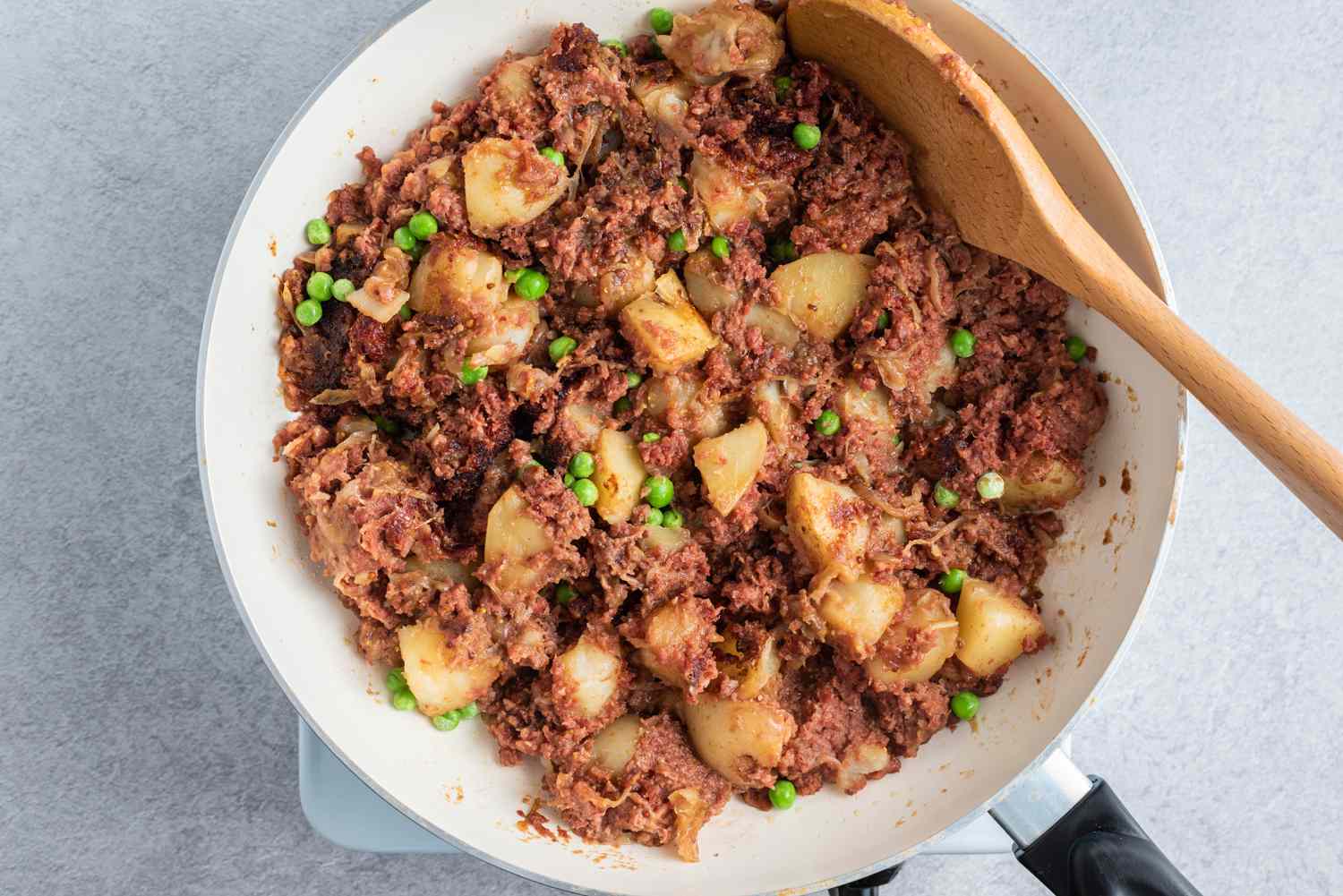 mixing corned beef with potatoes onions and peas in a large frying pan