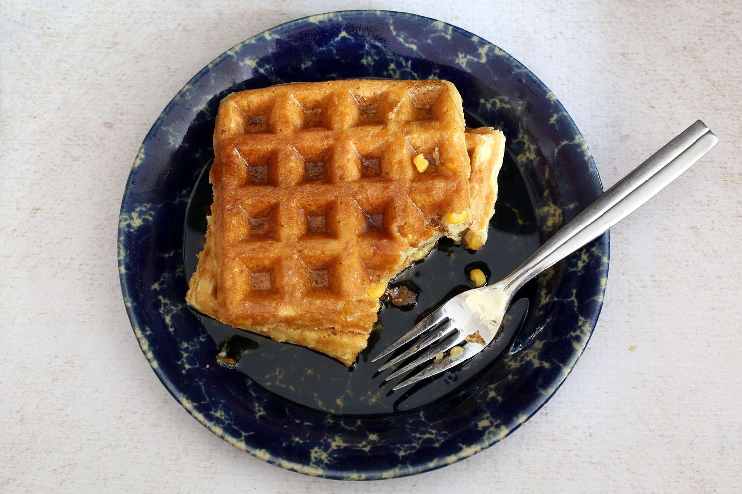 Two corn waffles on a blue plate with a waffle corner cut off by a fork