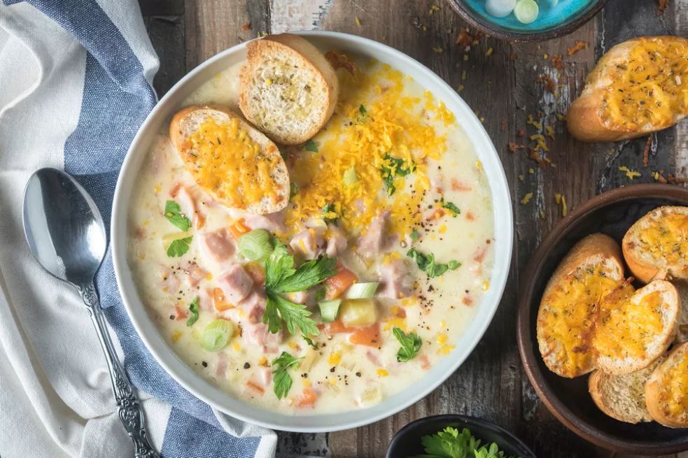 An overhead of a bowl full of creamy potato soup with ham and a side of cheese covered bread