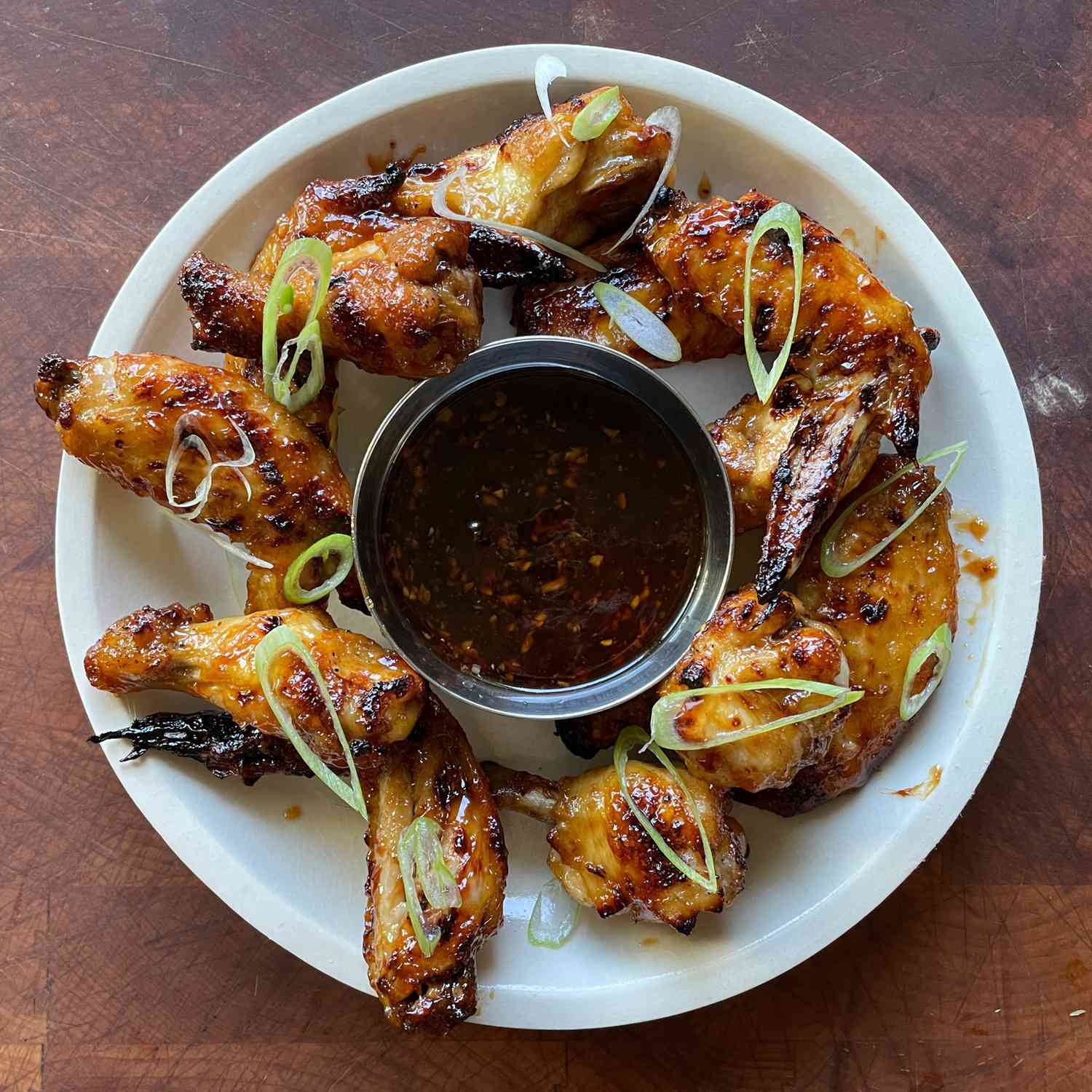 Deep golden brown chicken wings on a plate with a metal bowl of sauce in the center; garnished with green onion