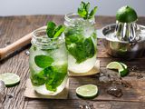 Two mojitos with mint added with a lime being juiced in the background