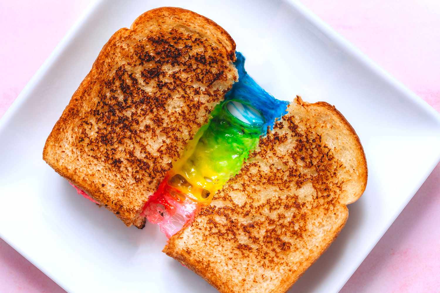 rainbow grilled cheese cut in half, on a plate, pulled apart to reveal blue, green, yellow, and pink cheese 