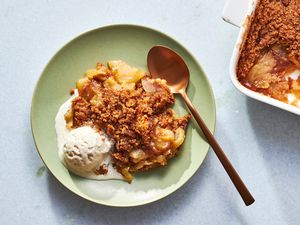 A serving of apple brown betty in a bowl, served with vanilla ice cream, with a baking dish of apple brown better on the side