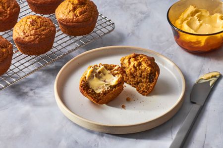 A pumpkin muffin, cut in half with butter, with a cooling rack of pumpkin muffins