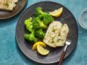 A plate with lemon-garlic baked cod, served with steamed broccoli, lemon wedges, and a glass of seltzer