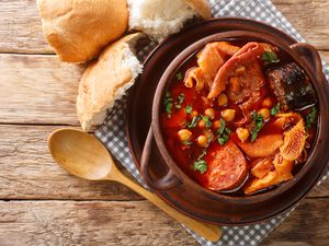 Wooden bowl full of made tripe, chorizo, blood sausage and chickpeas in a spicy sauce. There is a gray checkered napkin under the bowl and a wooden spoon next to the dish. Above the bowl there are broken up pieces of bread. 