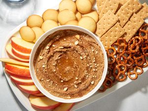 A bowl of snickerdoodle hummus garnished with maple syrup, crushed vanilla wafers, and cinnamon, served on a platter with slices apples, vanilla cookies, graham crackers, and pretzels