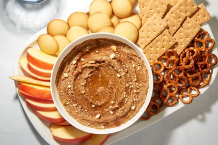 A bowl of snickerdoodle hummus garnished with maple syrup, crushed vanilla wafers, and cinnamon, served on a platter with slices apples, vanilla cookies, graham crackers, and pretzels