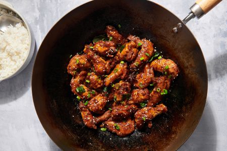 A wok of three cup chicken garnished with sesame seeds and scallions, served with a side of white rice