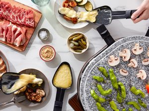 A raclette cheese party with a raclette grill, roasted vegetables, cured meats, pickles, and mustard