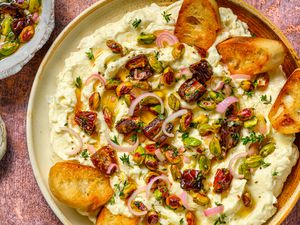 A platter of whipped brie topped with nuts, dates, pickled shallots, and fresh herbs served with toasted bread