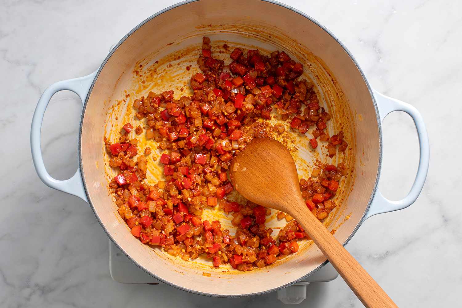 Onions and pepper with garlic, harissa, if using, paprika, cumin, and coriander in a pot on a burner, with a wooden spoon