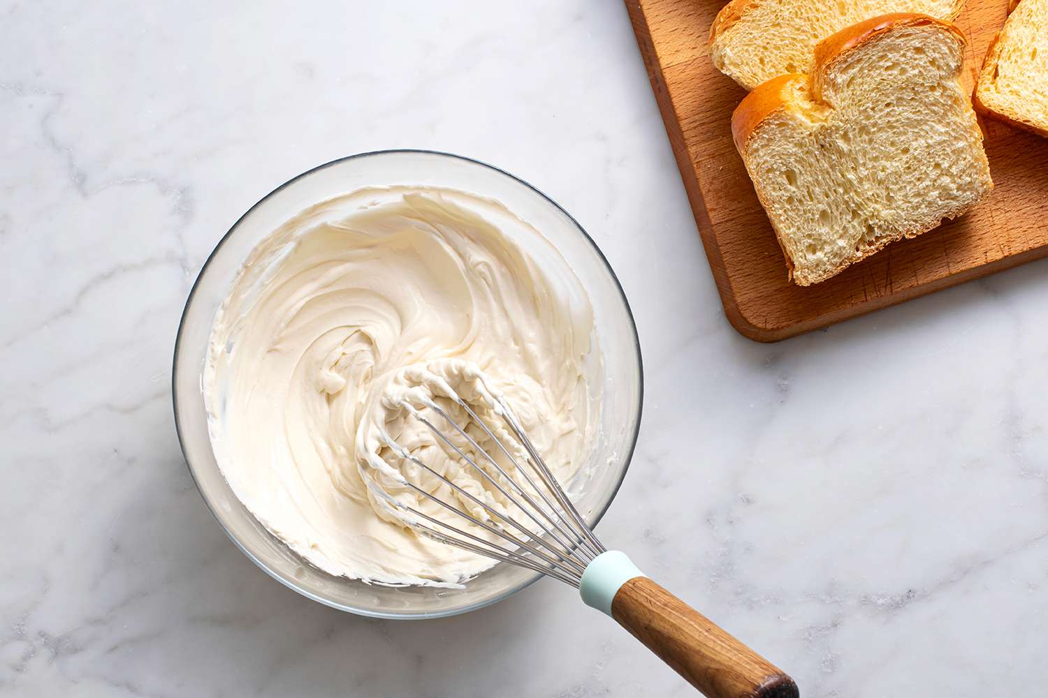 Bowl filled with cream cheese mixture and a whisk, slices of prepared bread on a cutting board.