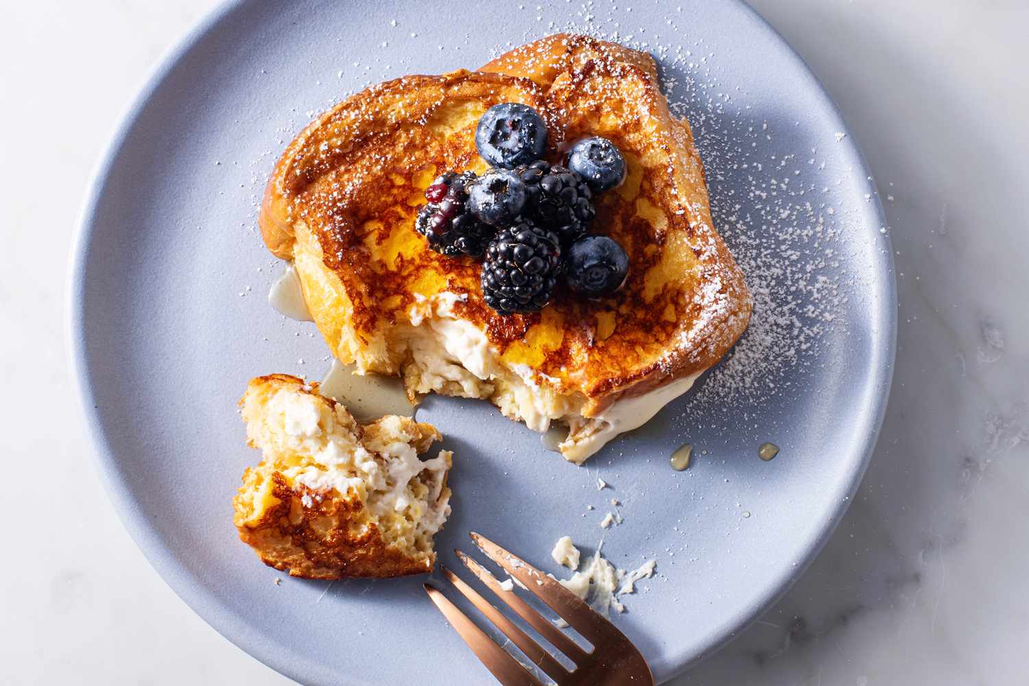 One piece of stuffed french toast on a plate topped with powdered sugar, blueberries and blackberries, maple syrup, with a piece cut out with a fork to reveal cream cheese filling.