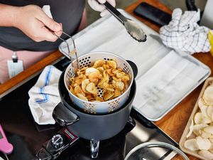 Hand lifting a strainer basket of potato chips out of a deep fryer while another hand holds a pair of tongs