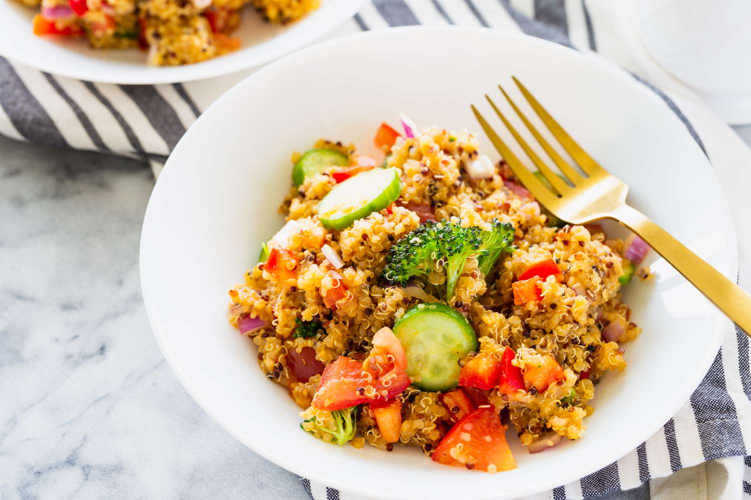 A Quinoa salad with broccoli, tomato and cucumber in a white bowl with a golden fork