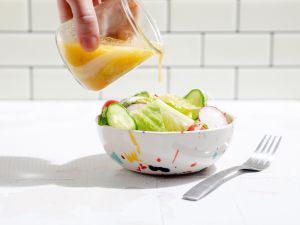 Salad dressing being poured from a small glass jar onto a romaine salad with sliced cucumbers and cherry tomatoes in a white bowl